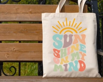 Peut inclure: Un sac fourre-tout en toile blanche avec un motif graphique coloré qui dit "SUN SHINE ON MY MIND". Le graphique est dans un style rétro avec un soleil jaune et des couleurs arc-en-ciel.