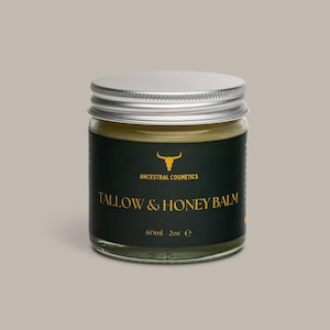 Op de afbeelding: Een glazen pot met een zilveren deksel met een balsem. De pot heeft een zwart etiket met gouden tekst met de tekst "TALLOW & HONEY BALM" en "ANCESTRAL COSMETICS". De pot bevat 60 ml balsem.