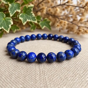 Peut inclure: Un bracelet élastique composé de perles rondes et polies en lapis-lazuli. Les perles sont d'un bleu profond et saturé, avec des inclusions naturelles de pyrite dorée et de calcite blanche visibles sur la surface des pierres.