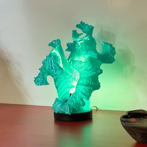 Può includere: Una lampada scultura astratta color turchese con una superficie strutturata, illuminata da una luce verde. La lampada è posizionata su una superficie rossa, accanto a una piccola ciotola scura. Il design della lampada ricorda una formazione corallina.