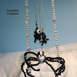 Peut inclure: Un mobile suspendu avec le texte Crystaline Creations, composé de deux brins verticaux de perles facettées transparentes avec des accents violets, deux chaînes argentées avec des grappes de perles noires et une araignée centrale en perles.