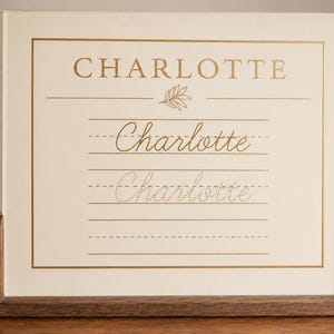 May include: A rectangular off-white sign with gold foil lettering and lines, held in a dark wood stand. The text reads CHARLOTTE in block capitals, followed by the name Charlotte in cursive script and a dotted line version for practice.