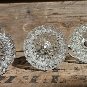 May include: Four clear glass cabinet knobs featuring a faceted, diamond-cut pattern. Each knob has a circular face with a central metal screw attachment visible in the middle, reflecting light through the textured, transparent glass surface.