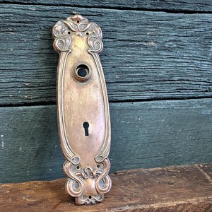 May include: An antique brass door plate with ornate scrollwork and a keyhole. The plate has a tarnished finish, showing its age. The design includes a central oval shape with decorative elements at the top and bottom. The door plate is approximately 7 inches long.