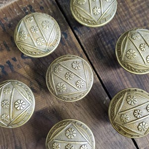 May include: Seven antique brass buttons with detailed geometric and floral patterns are arranged on a weathered wooden surface. The buttons have a domed shape and a warm, metallic finish, showcasing intricate designs.