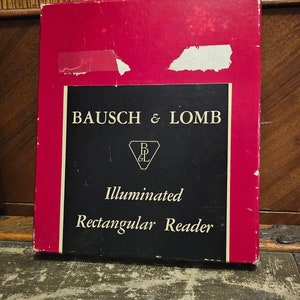 May include: A red and black box with the text "BAUSCH & LOMB" and "Illuminated Rectangular Reader". The box has a diamond-shaped logo with the letters "B&L" inside.