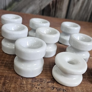May include: A collection of small, white ceramic candle holders. Each holder has a circular top and base, connected by a narrower, cylindrical middle section. The holders are arranged on a rustic, wooden surface.