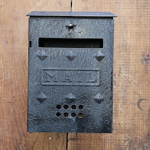 May include: Black metal mailbox with a rectangular slot for mail and the word "MAIL" embossed on the front. The mailbox has a textured surface and is decorated with diamond-shaped studs. It is mounted on a wooden surface.