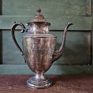 May include: A silver teapot with a rounded body and a decorative handle. The teapot has a tarnished finish and sits on a wooden surface.