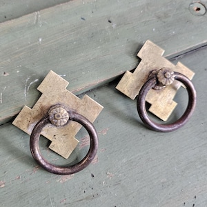 May include: Two antique brass drawer pulls with a dark ring handle and a decorative floral center. The pulls are mounted on a square, cross-shaped brass backplate. The background is a distressed, painted green surface.