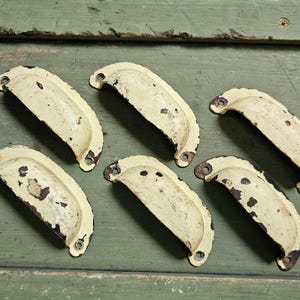 May include: A set of six semi-circular metal drawer pulls with a distressed cream-colored finish. Each handle features a curved grip and two mounting holes at the ends, showing significant dark brown paint wear and oxidation across the surfaces.