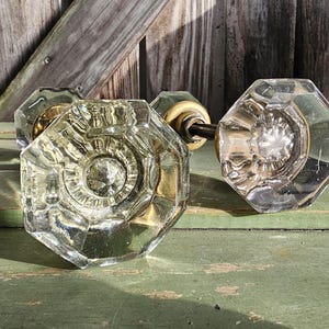 May include: A pair of clear glass door knobs featuring an octagonal shape with faceted edges. Each knob is attached to a brass-toned metal base and connected by a central metal spindle, showcasing a reflective and transparent design.