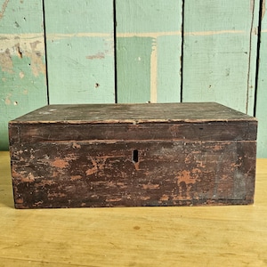 May include: A rectangular wooden box with a dark brown, weathered finish and visible signs of wear. The box features a hinged lid and a central vertical keyhole on the front panel. The wood grain is visible throughout the surface of the box.