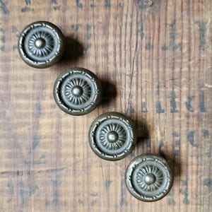 May include: Four circular metal cabinet knobs arranged in a diagonal line. Each knob features a dark bronze finish with a central raised dome surrounded by a radiating sunburst pattern and a textured outer rim with small repeating indentations.