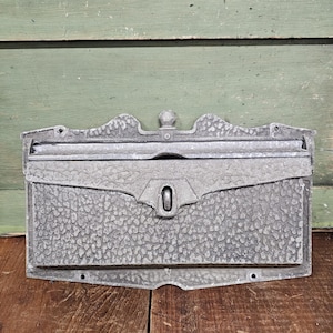 May include: A weathered, gray metal mailbox with a textured surface and a decorative top. The mailbox has a keyhole and a slot for mail. The background is a green wooden wall and a wooden surface.