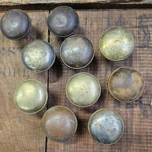May include: A collection of nine round metal cabinet knobs featuring various weathered finishes including tarnished brass, aged copper, and dark oxidized steel. Each knob has a smooth, domed surface with visible signs of wear, patina, and texture.