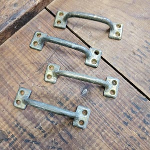May include: Four vintage brass drawer pulls with a curved handle design. Each pull has two rectangular mounting plates with two screw holes each. The pulls are arranged on a weathered wooden surface.