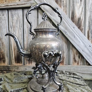 May include: An antique silver-plated tea kettle with ornate detailing. The kettle features a curved spout, a decorative handle, and a lid with a finial. It sits on a matching stand with elaborate, stylized legs. The background is a rustic wooden wall.