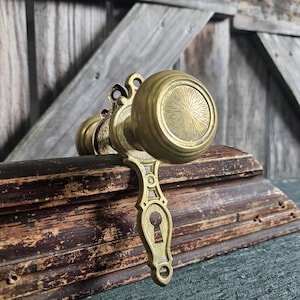 May include: Antique brass door knob set with a decorative backplate and keyhole. The knob has a circular design with a sunburst pattern. The set is placed on a weathered wooden surface, with a rustic wooden background.