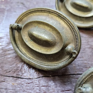 May include: An oval-shaped brass drawer pull featuring a central raised medallion with a sunburst pattern. The hardware has a beaded border and a swinging bail handle attached to two small circular mounts on the sides, showing a weathered patina.