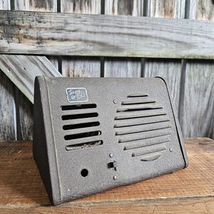 May include: A vintage, brown metal speaker with horizontal and vertical vents. The speaker has a small switch and a label that reads "Tavany & Filkie". The speaker is angled on a wooden surface.