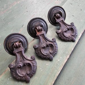 May include: Three antique bronze drawer pulls with ornate, scrollwork designs. Each pull features a circular backplate and a decorative handle. The pulls are arranged on a weathered, green-painted wooden surface.