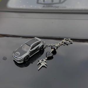 May include: A silver and black miniature car keychain with a black roof and hood. The keychain includes a chain, black beads, a black heart, and a silver star and cross charm.