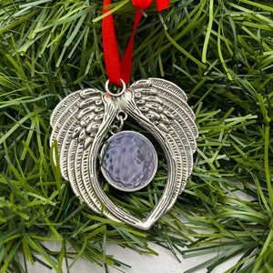 May include: Silver angel wing ornament with a blue and purple glass center. The ornament is hanging from a red ribbon.