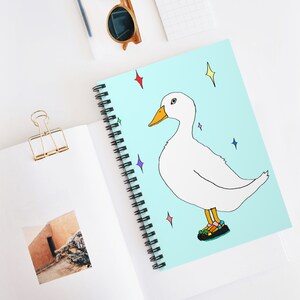 May include: Spiral-bound notebook with a light blue cover featuring a cartoon duck wearing sandals. The duck is white with a yellow beak and is surrounded by colorful star-shaped sparkles. The notebook has a black spiral binding.