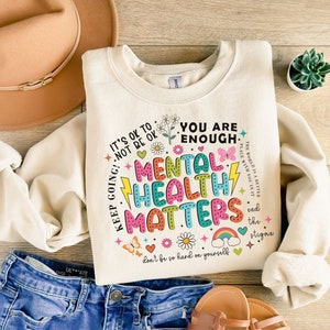 May include: A cream colored sweatshirt with a colorful graphic print that says "It's ok to not be ok. Keep going! You are enough. Mental Health Matters. Don't be so hard on yourself. The world is a better place with you in it. End the stigma."