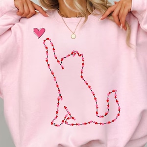 May include: A light pink sweatshirt featuring a cat silhouette outlined with red and pink hearts. A single pink heart floats above the cat's head. The sweatshirt has a crew neck and long sleeves.