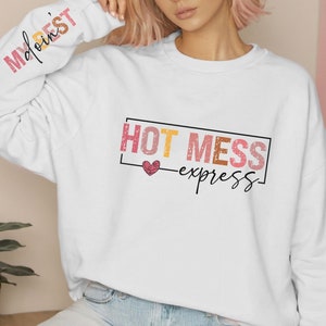 Może przedstawiać: Biała bluza z nadrukiem graficznym, na kt&oacute;rym widnieje napis "HOT MESS express" w r&oacute;żowych, pomarańczowych i ż&oacute;łtych literach z małym r&oacute;żowym sercem.