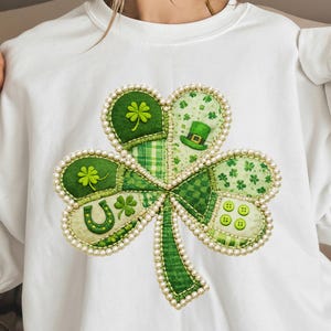 Può includere: Felpa bianca con un grande e dettagliato design a trifoglio. Il trifoglio è composto da vari motivi in tessuto verde, tra cui trifogli, un cappello da folletto e un ferro di cavallo. I bordi sono bordati con un bordo perlato, creando un tema festivo per il giorno di San Patrizio.