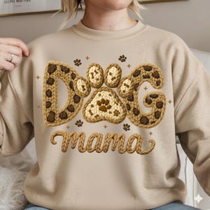 May include: Beige sweatshirt with the word "DOG" in a brown leopard print design and a paw print in the center. The word "mama" is written in a cursive font below the paw print. Small paw prints are scattered around the design.