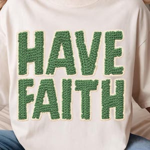 May include: Cream-colored sweatshirt with the words "HAVE FAITH" in large, textured, green letters. The letters have a light beige border, creating a bold contrast against the shirt's neutral background. The sweatshirt has a relaxed fit.