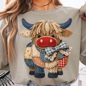 May include: A gray sweatshirt featuring a patchwork cow design. The cow has blue denim horns, a red nose, and a plaid bow. The cow's body is made of various fabric patterns, including floral and gingham.