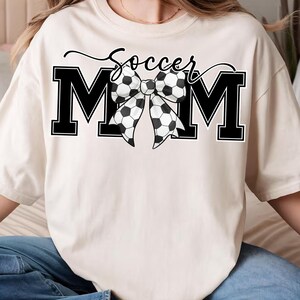 May include: Cream-colored t-shirt with the text "Soccer Mom" in black script above large black block letters spelling "MOM". A bow made of soccer balls is centered between the letters.