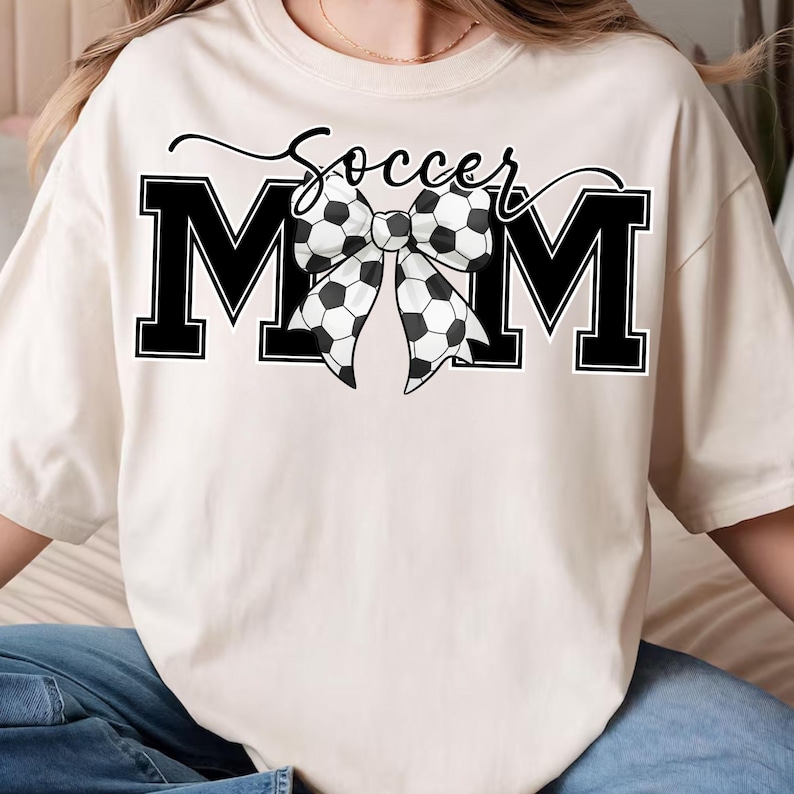 May include: Cream-colored t-shirt with the text "Soccer Mom" in black script above large black block letters spelling "MOM". A bow made of soccer balls is centered between the letters.