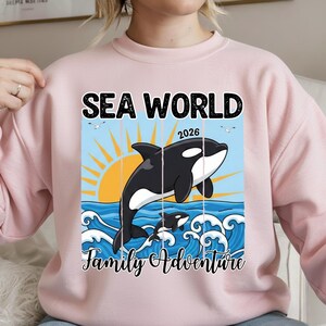 K&ouml;nnte beinhalten: Rosa Sweatshirt mit einer Grafik, die einen schwarz-wei&szlig;en Orca-Wal zeigt, der aus blauen Wellen unter einer gelben Sonne springt. Der Text "SEA WORLD 2026" und "Family Adventure" sind &uuml;ber und unter der Wal-Illustration abgebildet.