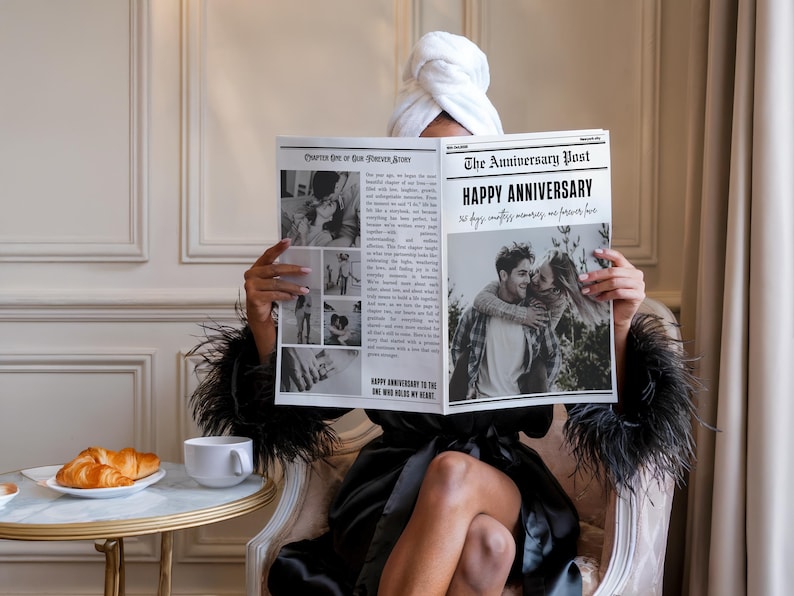 May include: A person in a black robe and a white towel on their head is holding a newspaper with the headline "HAPPY ANNIVERSARY". A white teacup and croissants are on a small table.