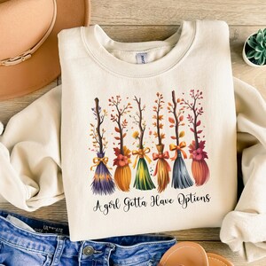 May include: Cream-colored sweatshirt featuring a design of six colorful broomsticks with autumn foliage and bows. The text "A girl Gotta Have Options" is printed below the brooms. The sweatshirt is paired with blue jeans and a brown hat.
