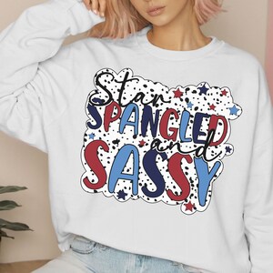 Puede incluir: Sudadera blanca con el texto "Star Spangled and Sassy" en rojo, blanco y azul. El diseño incluye estrellas y lunares negros. Una prenda superior informal de temática patriótica.