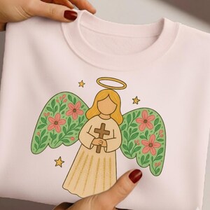 May include: A light pink sweatshirt featuring a cartoon angel design. The angel has blonde hair, a gold halo, and holds a wooden cross. The angel's wings are green with pink flowers. Three gold stars are also featured in the design.