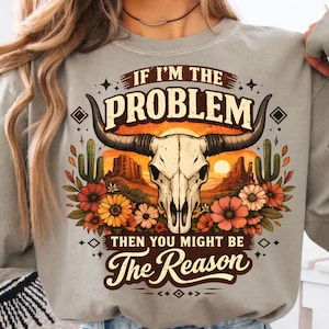 Può includere: Felpa grigio chiaro con una grafica di un teschio di toro con corna, ambientata in un tramonto nel deserto. Il testo recita "IF I'M THE PROBLEM THEN YOU MIGHT BE The Reason". Il design include cactus e fiori.