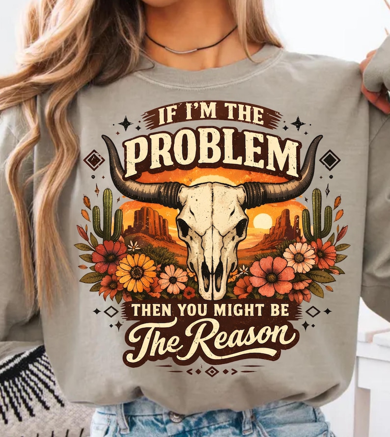 May include: A light gray sweatshirt featuring a graphic of a bull skull with large horns, set against a desert sunset scene. The text reads "IF I'M THE PROBLEM THEN YOU MIGHT BE The Reason". The design includes cacti and flowers.