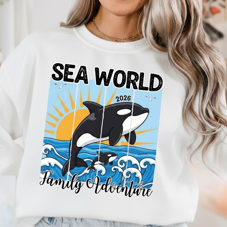K&ouml;nnte beinhalten: Wei&szlig;es Sweatshirt mit einer Grafik, die einen springenden Orca-Wal, einen kleineren Orca und den Text "SEA WORLD 2026 Family Adventure" &uuml;ber den Meereswellen und einer untergehenden Sonne zeigt.