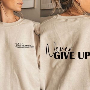 Op de afbeelding: Een beige sweatshirt met de tekst "you have the power to change your story" op de voorkant en "Never Give Up!" op de achterkant.