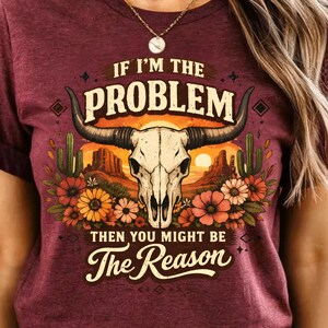 May include: Burgundy t-shirt with a graphic design featuring a bull skull, cacti, and flowers. The text reads "IF I'M THE PROBLEM THEN YOU MIGHT BE The Reason". The design has a desert sunset backdrop.