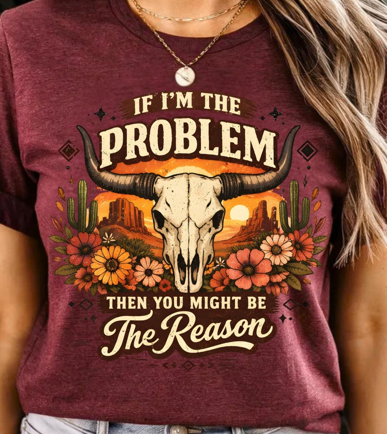 May include: Burgundy t-shirt with a graphic design featuring a bull skull, cacti, and flowers. The text reads "IF I'M THE PROBLEM THEN YOU MIGHT BE The Reason". The design has a desert sunset backdrop.