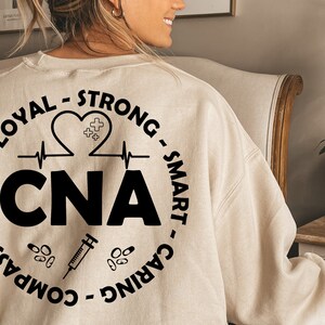 May include: A beige sweatshirt with a black circle design on the back. The design includes the words "CNA", "Loyal", "Strong", "Smart", "Caring", "Compassionate", and "Heart".